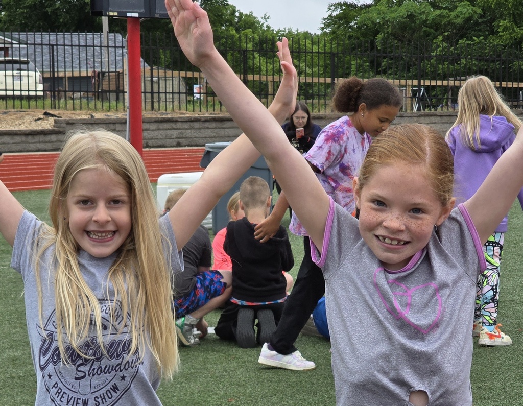 Two young girls with raised arms, smiling. Behind them, other children sit on the ground. A black fence borders the grassy area.