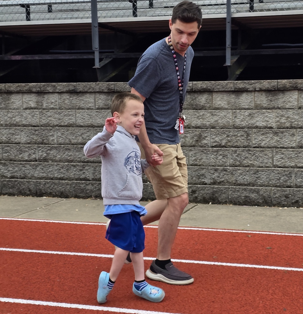 An adult and a child walk on a red track. The child has one arm raised. Behind them is a stone wall.
