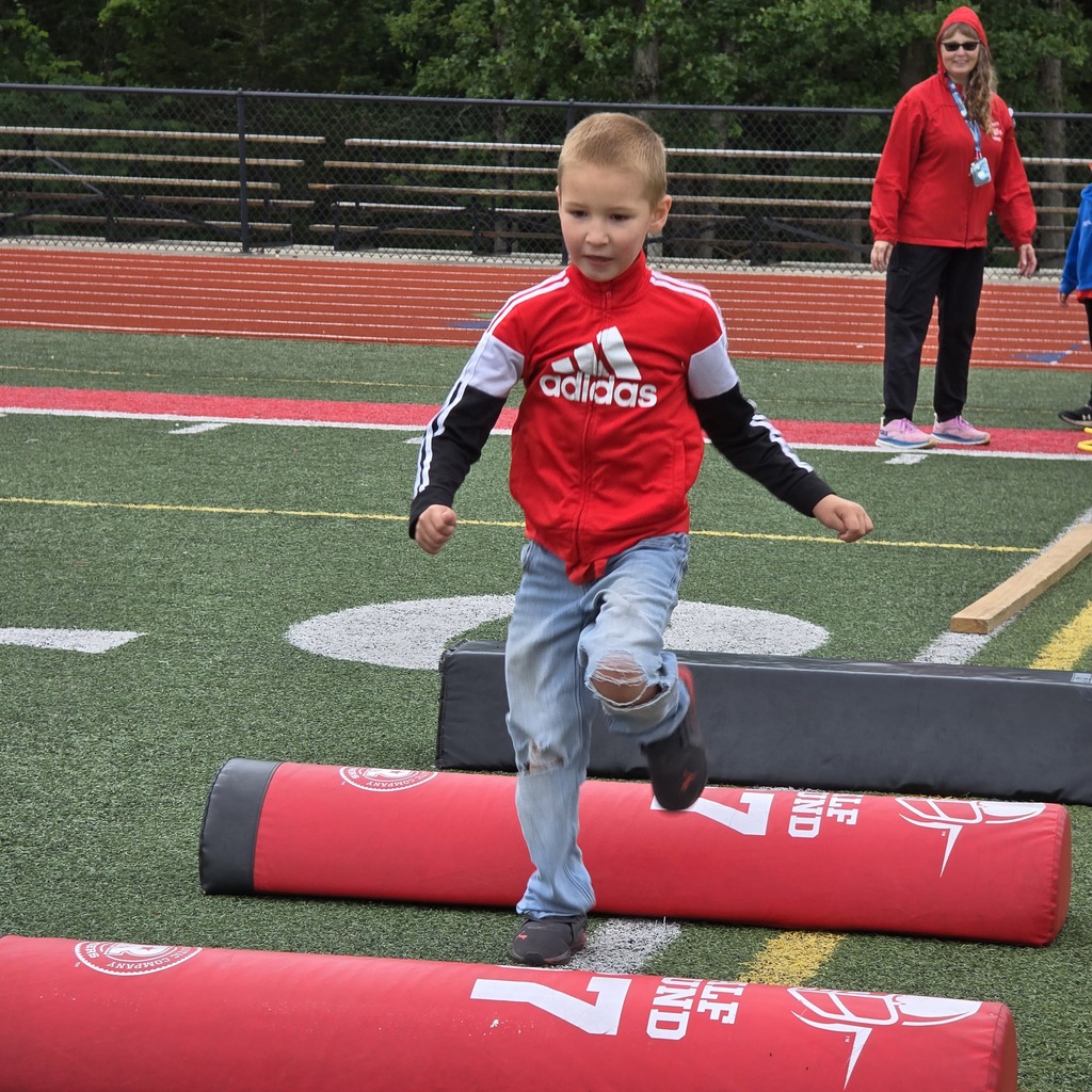 A young boy runs over hurdles outdoors on a field. A woman in red stands behind him.