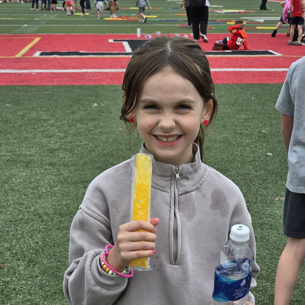 Girl in gray sweatshirt holds yellow popsicle and smiles. In background, children play on a field with red and green zones.