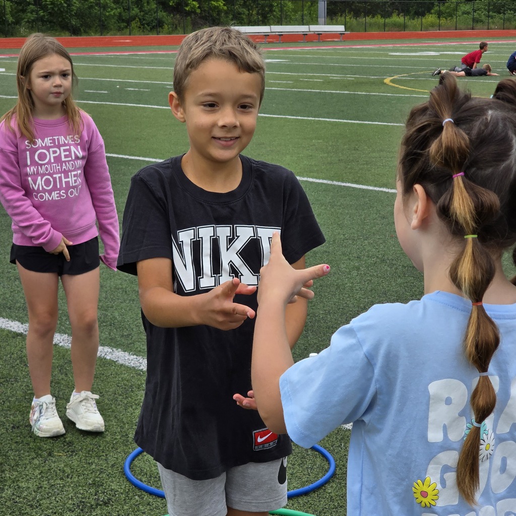 Children stand on a sports field, one talking to another. Two hula hoops lie on the ground.