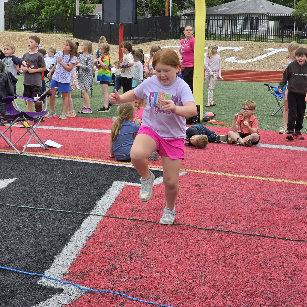 Children line up at a track, one girl leads in pink shorts. Other children sit or stand nearby.