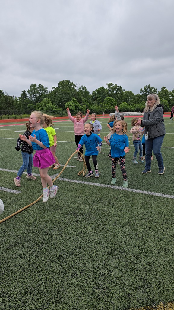 Kids in sports gear play a tug-of-war on a field with trees and cloudy sky in the background.