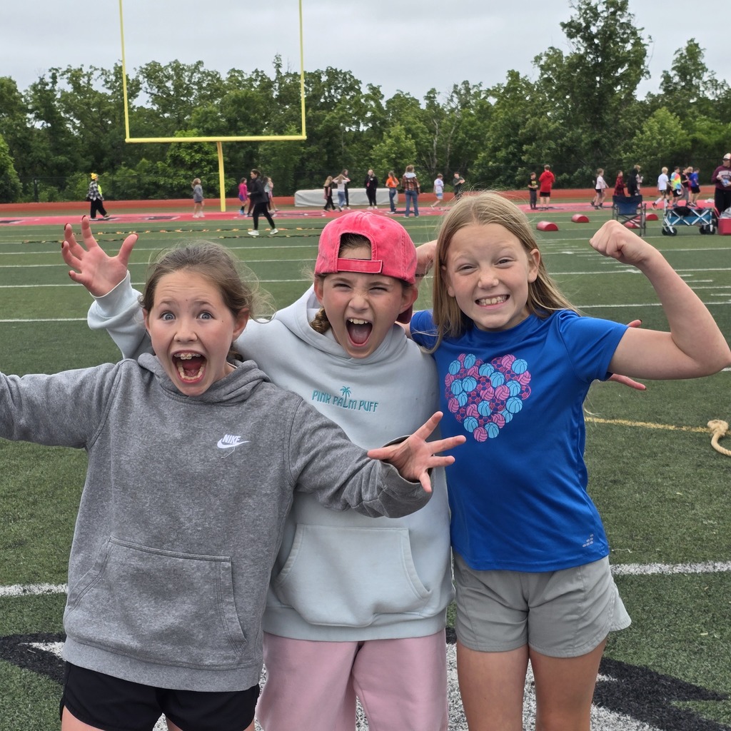 Three young girls pose for a photo on a football field with others in the background.