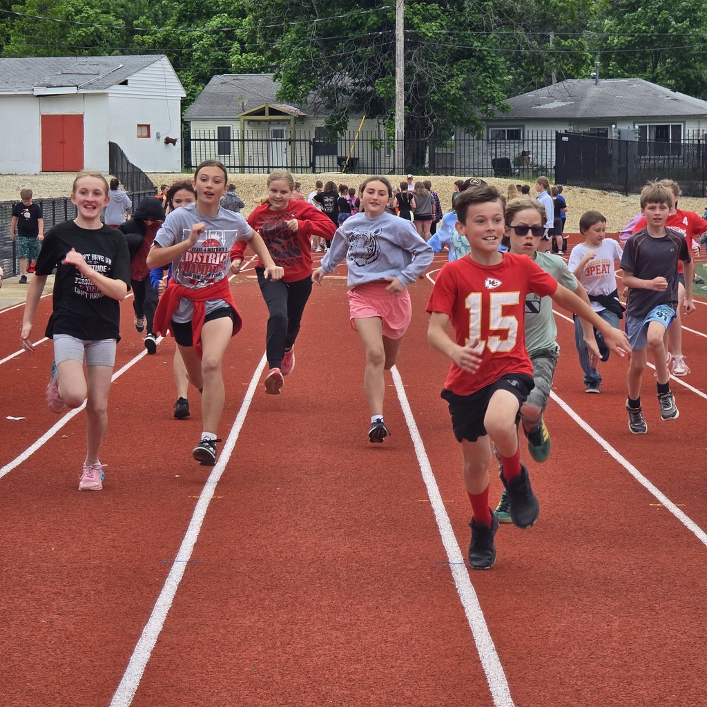 Multiple children in sports gear sprint on a track, with buildings and trees in the background.