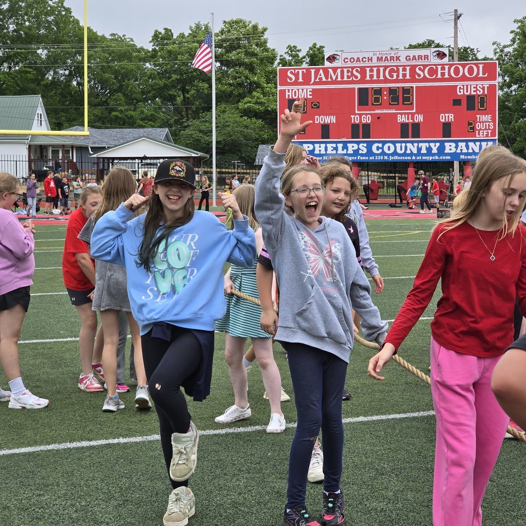 A group of young girls and others dance on a football field with a scoreboard in the background.