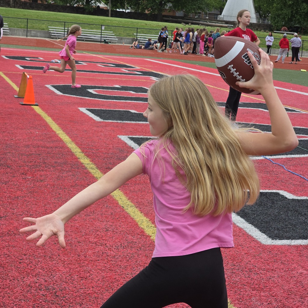A young girl throwing a football on a red field. Other players and spectators are in the background.