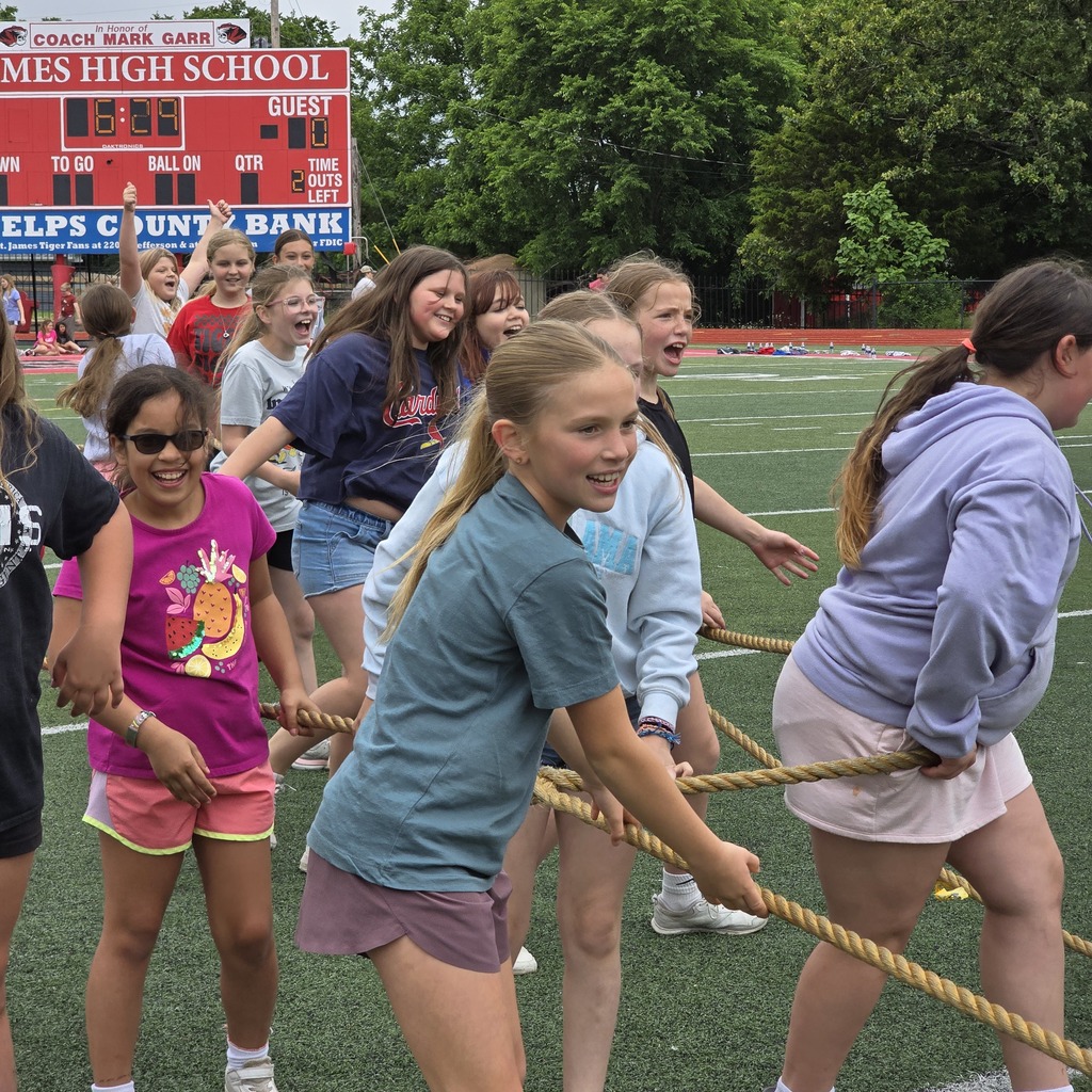 Students participate in a rope challenge at a sports field. A red banner behind them reads "James High School."