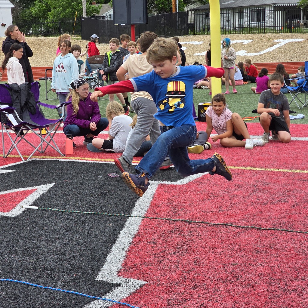 Children running and jumping on a track with red and black markings, some seated in chairs.