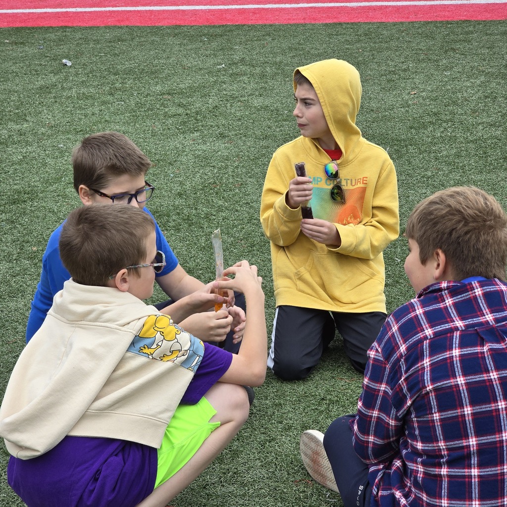 Four boys in various colored clothing are sitting on a grass field, engaging with objects. One holds a bottle.