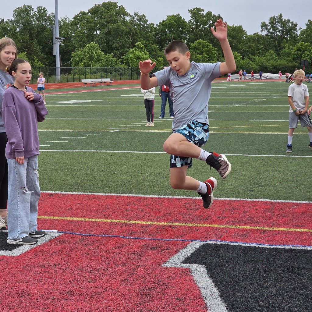 A boy jumps into a sand pit on a field, wearing shorts and sneakers. Behind him, others stand on the field.