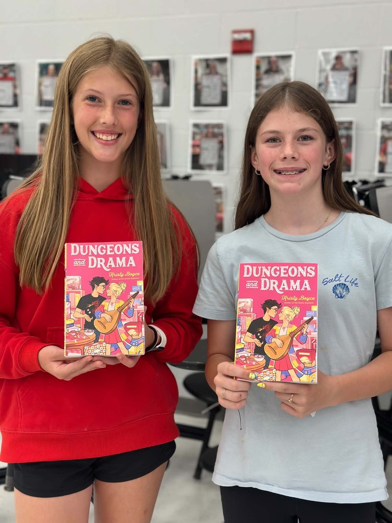 Two girls stand in a room, smiling and holding up books. One holds a pink "Dungeons and Drama" book; the other holds a blue one.