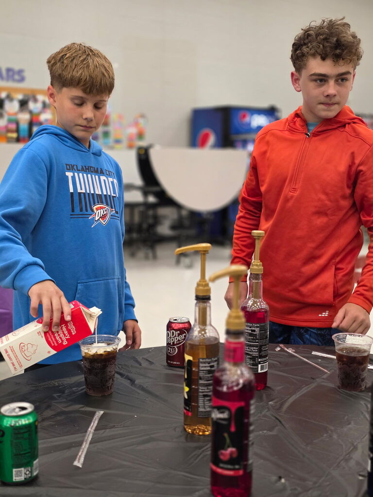 Two boys standing at a table with various drinks, one pours a bottle into a cup.