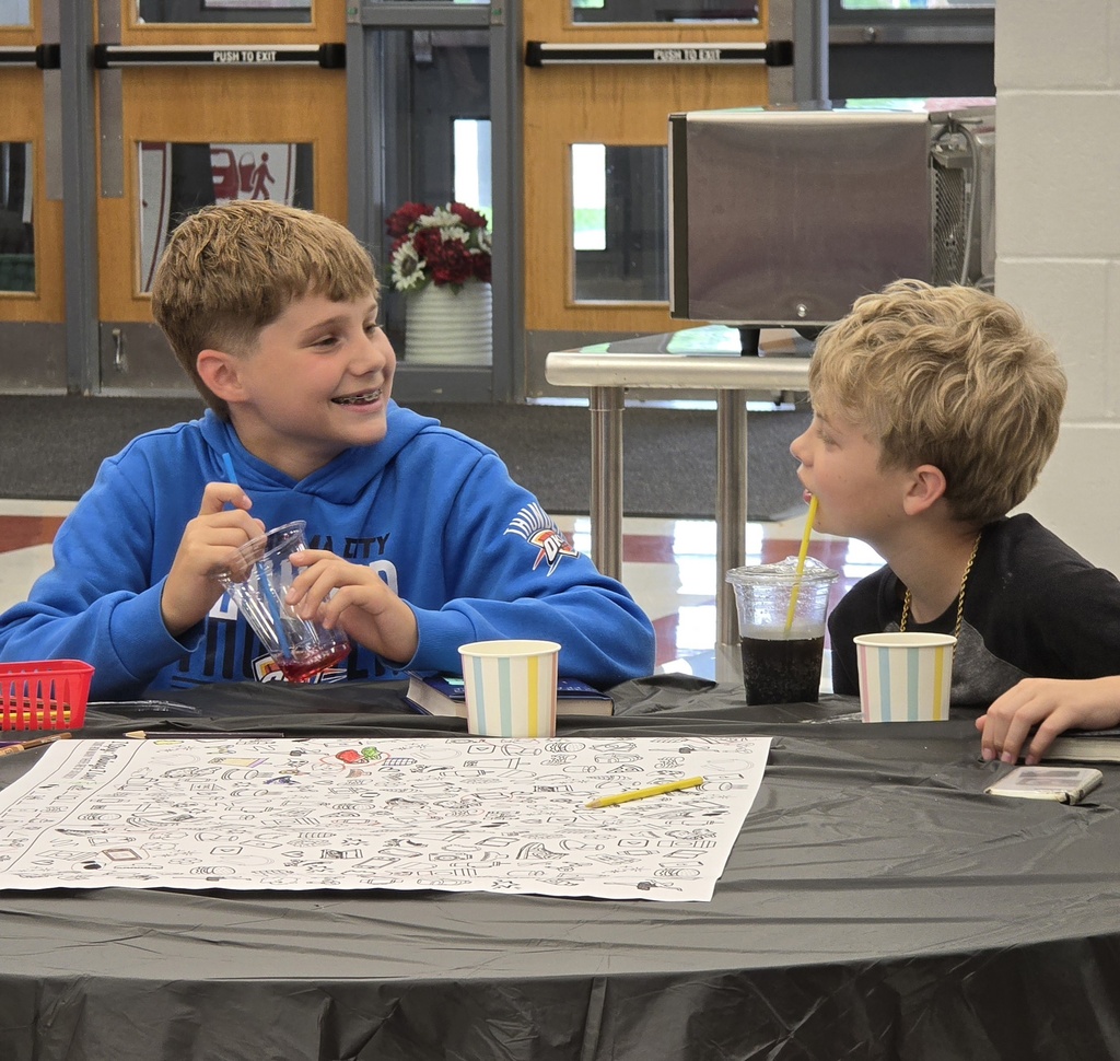 Two boys seated at a table, one smiling, the other holding a drink. A flower arrangement and a monitor behind them.