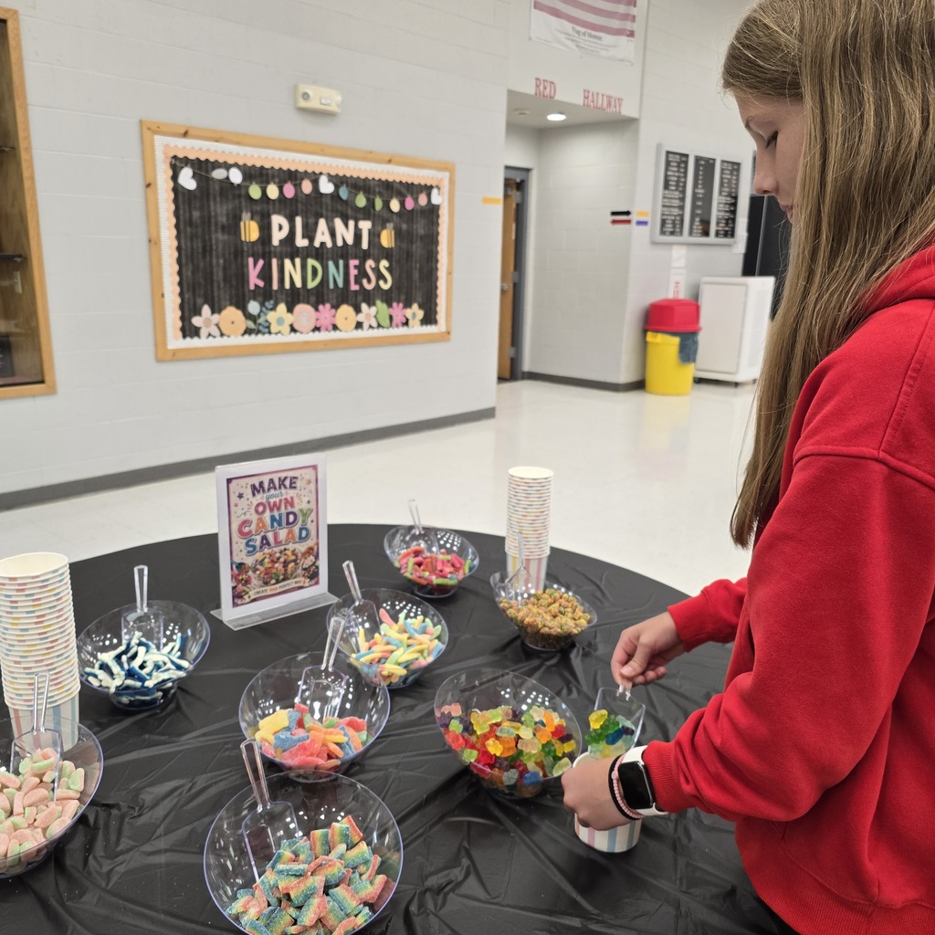 Person in a red hoodie picking up candies from bowls at a table. Behind them, a board displays "Plant Kindness".