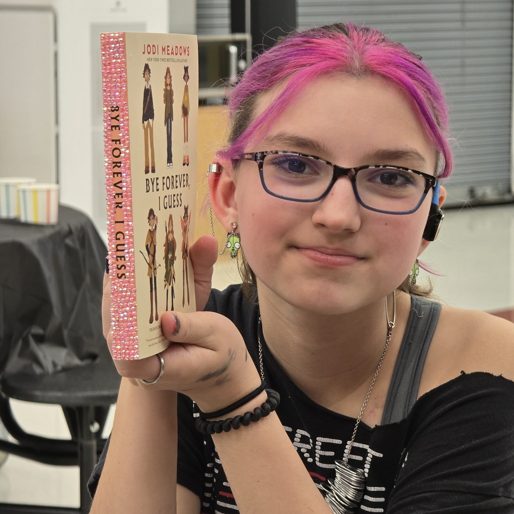 Person with pink hair holding a book titled "Bye Bye Unicorn" in a classroom.