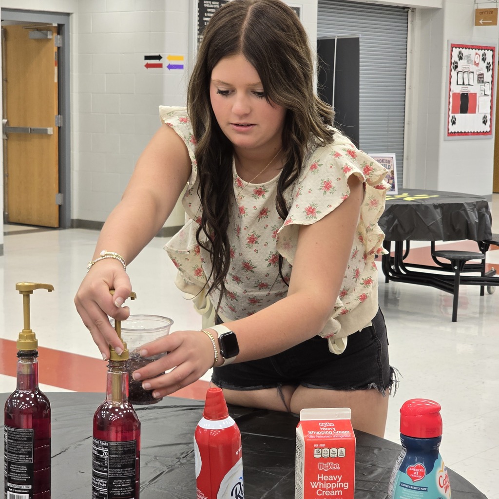 Woman standing at a table pours a red liquid into a cup from a bottle.