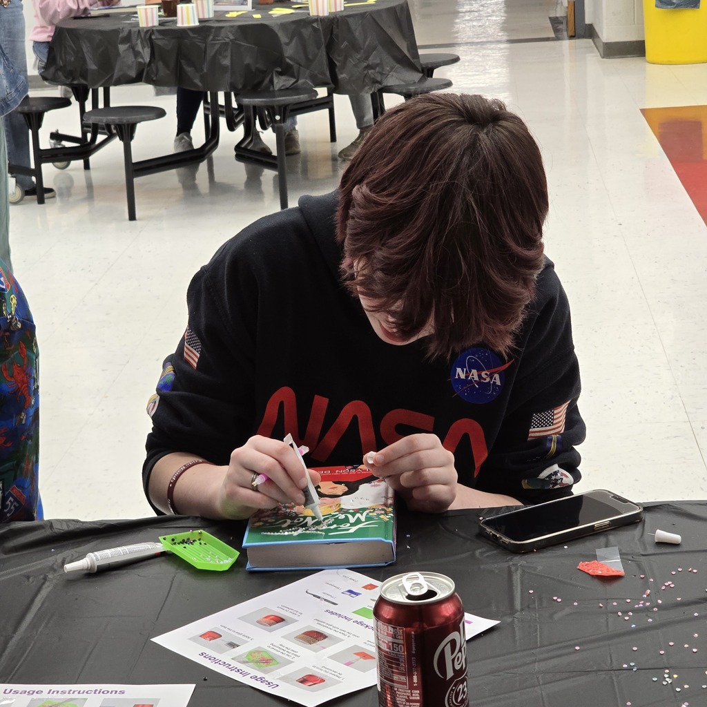 A person works at a table with a book, pen, phone, and other items. They wear a NASA shirt.