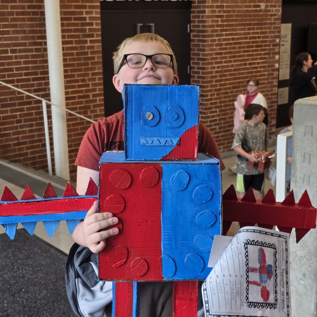 A child wearing glasses stands with a cardboard robot sculpture. The sculpture is blue, red, and white.