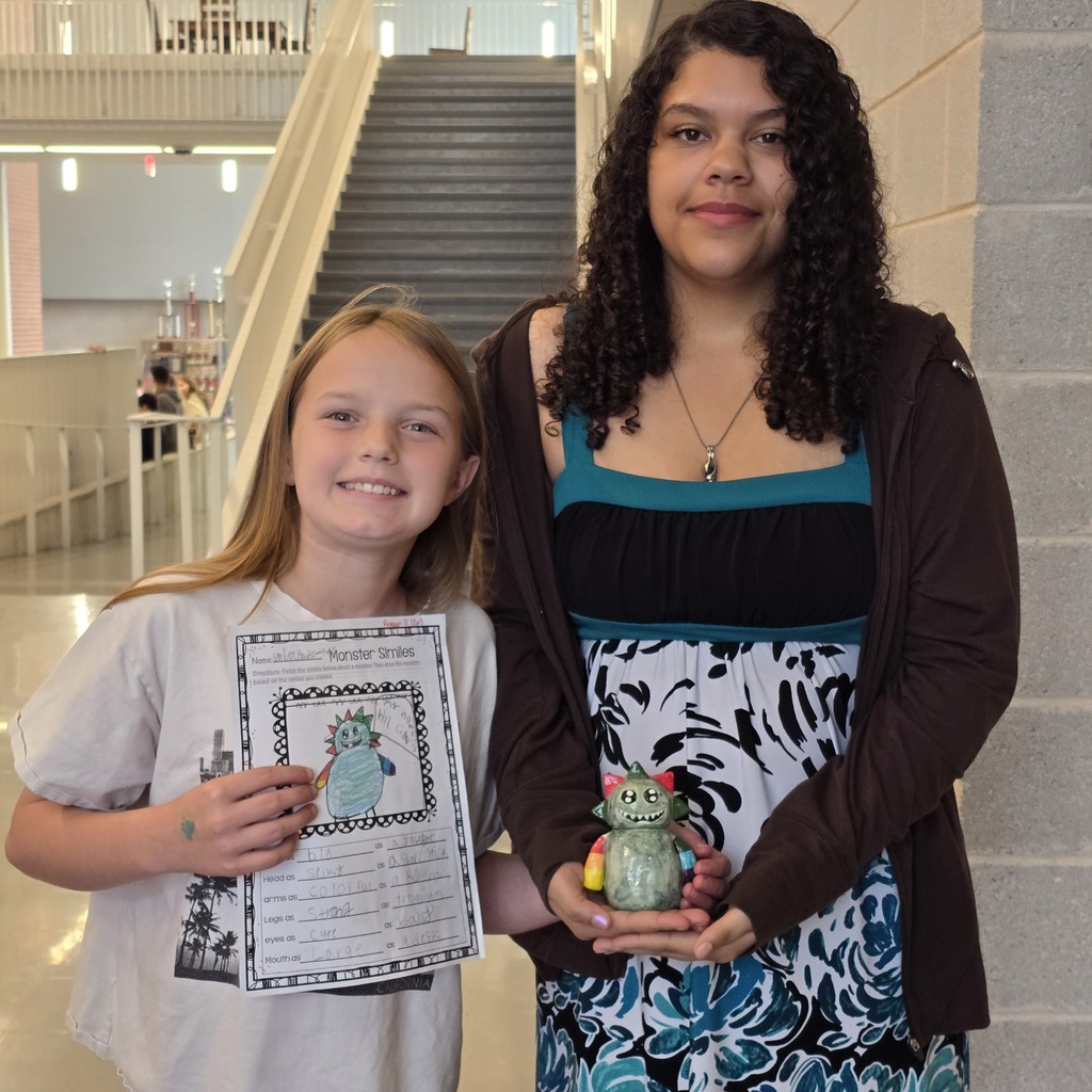 Two girls stand in a hallway, one holds a drawing, the other a small figurine. They smile and pose.