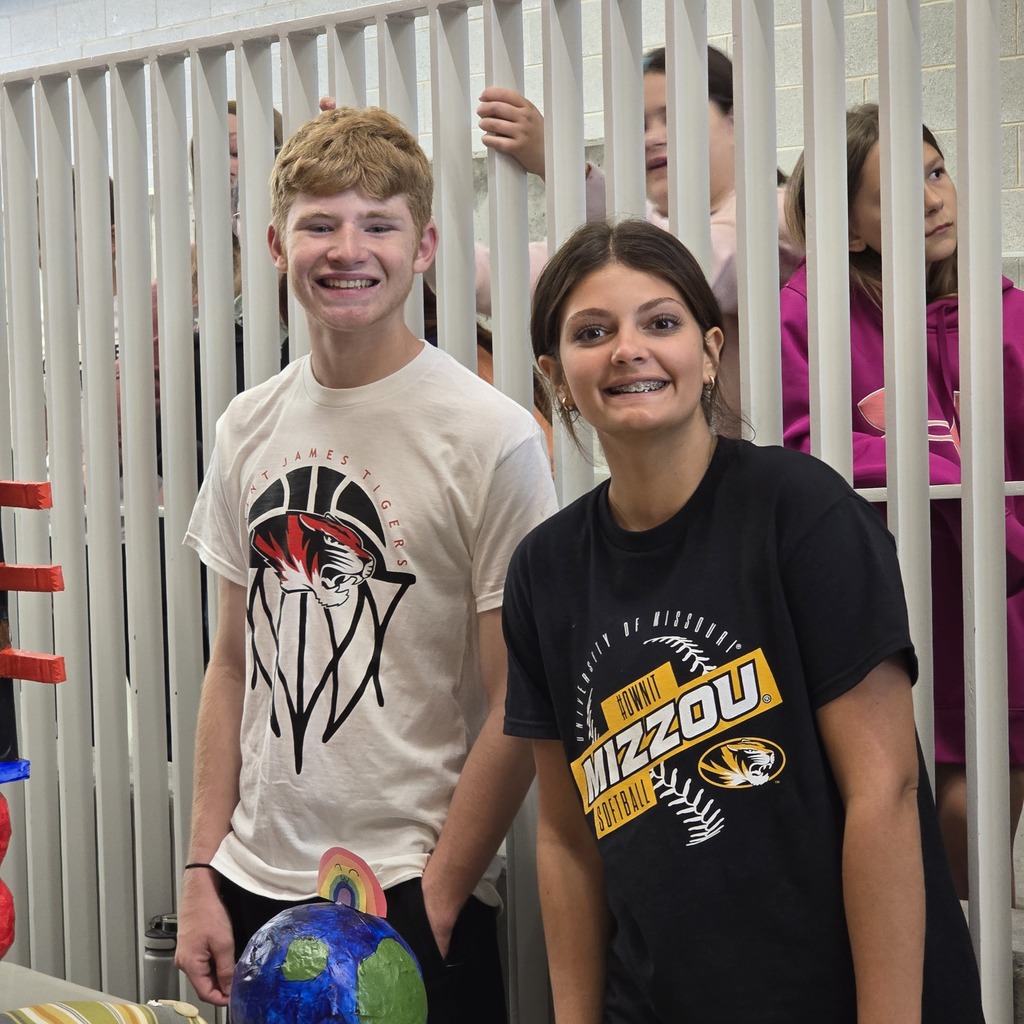 Two smiling individuals in a room, one in a white shirt and the other in a black shirt.