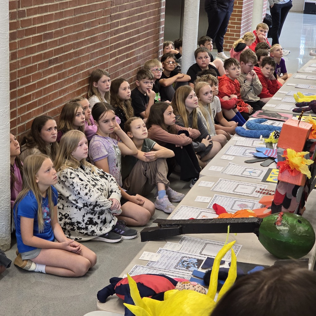 Children seated on the floor near a brick wall, some wearing glasses, one holding a watermelon.