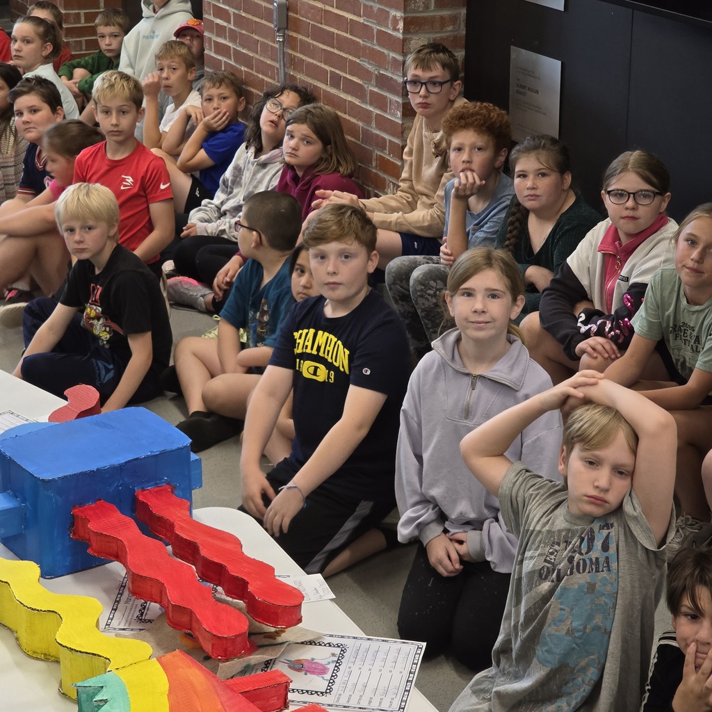 Group of students seated, some looking at colorful train models on a table. Brick wall in background.