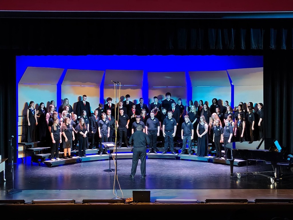 A choir performs on stage, illuminated by blue stage lighting. The conductor stands center, with musicians on either side.