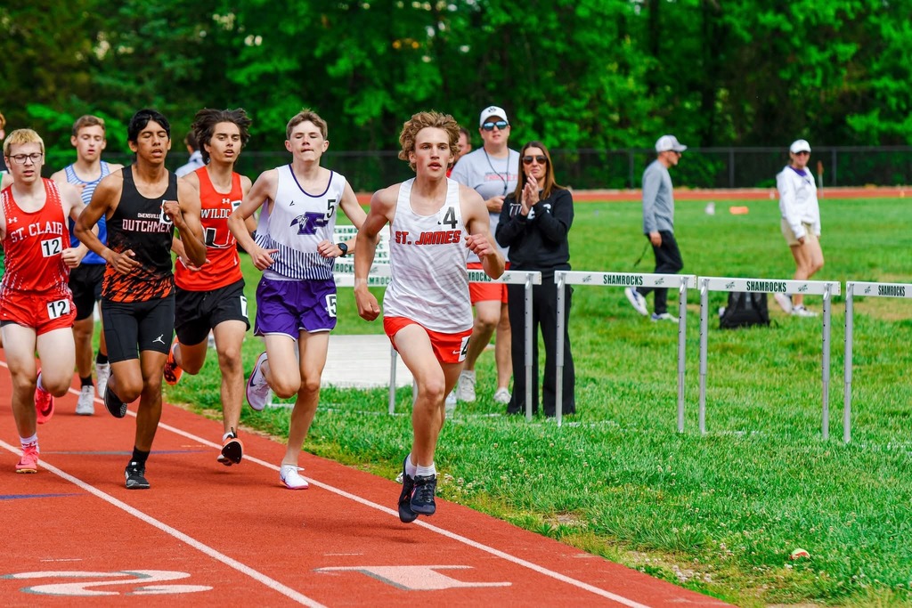 Runners compete on a track with hurdles. Participants wear numbered bibs. Spectators watch from the sidelines.