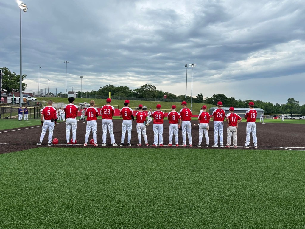 Eleven players in red uniforms stand in a row on a grassy field, facing the camera.