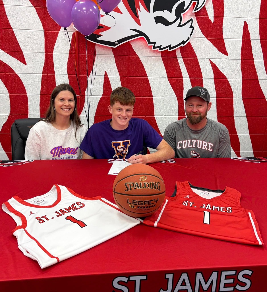 Three individuals sit at a table with a basketball, jersey, and balloons. A woman and boy wear white and blue, and a man in a gray shirt.