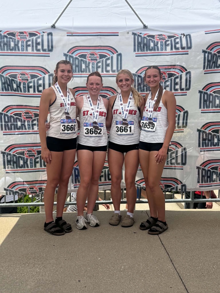 Four young women with medals stand together. They wear matching white shirts and black shorts. Behind them, a white banner says "TRACK & FIELD".