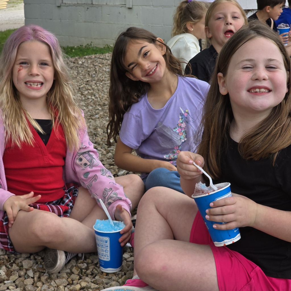 Group of girls sitting on gravel outdoors. Each holds a cup, two with straws. Smiling, one in pink jacket, one in black, one in purple.