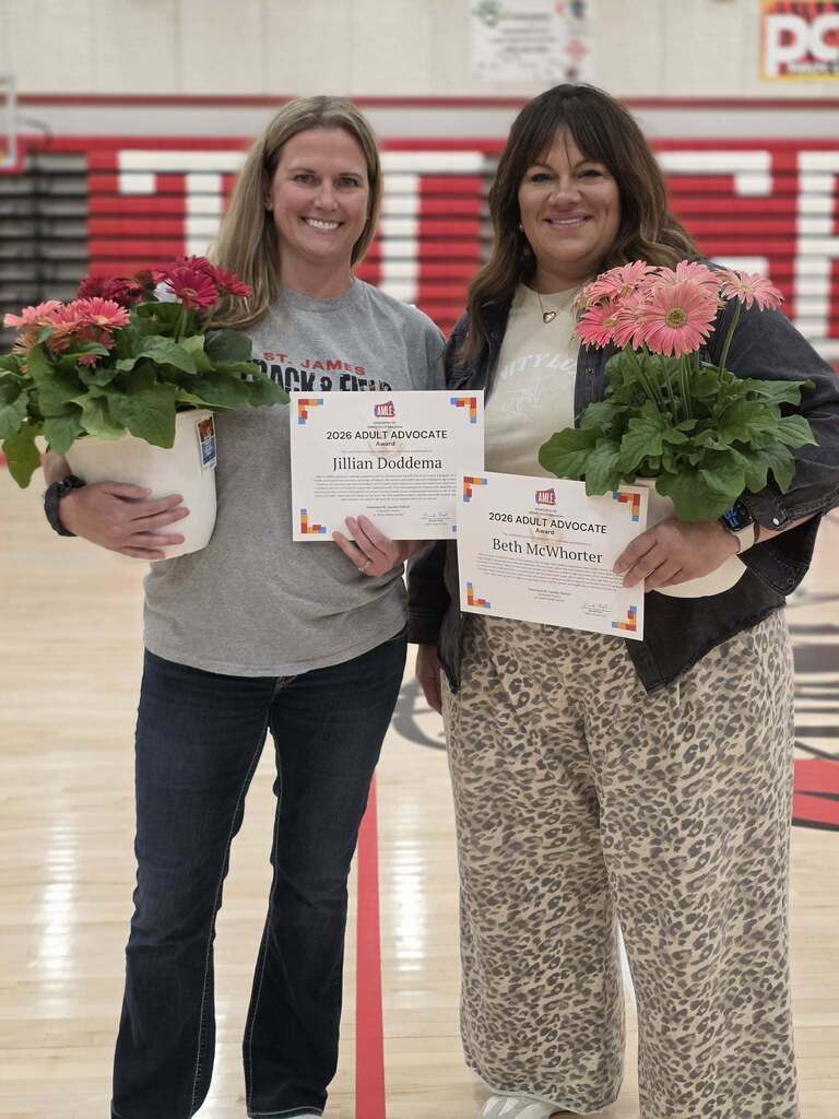 Two women standing in a gym holding certificates and flower pots, one woman holding a white certificate.