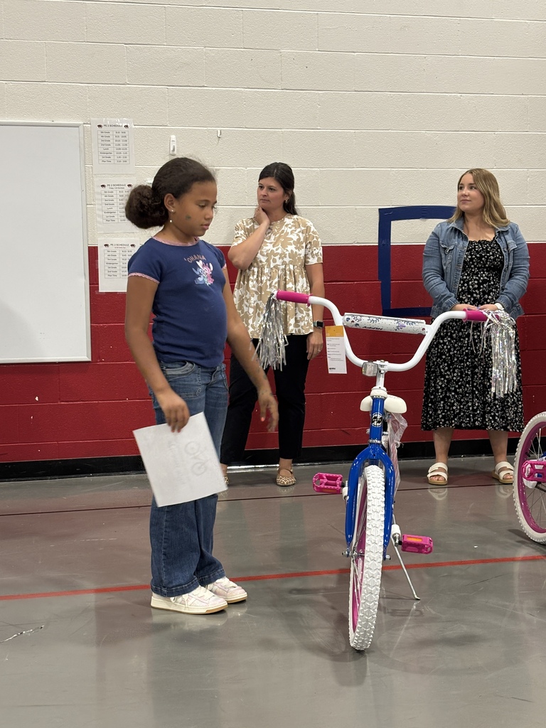 A girl in a blue shirt holds a paper while standing next to a white bicycle. Behind her, two women stand, one wearing a blue jacket and the other a black dress.