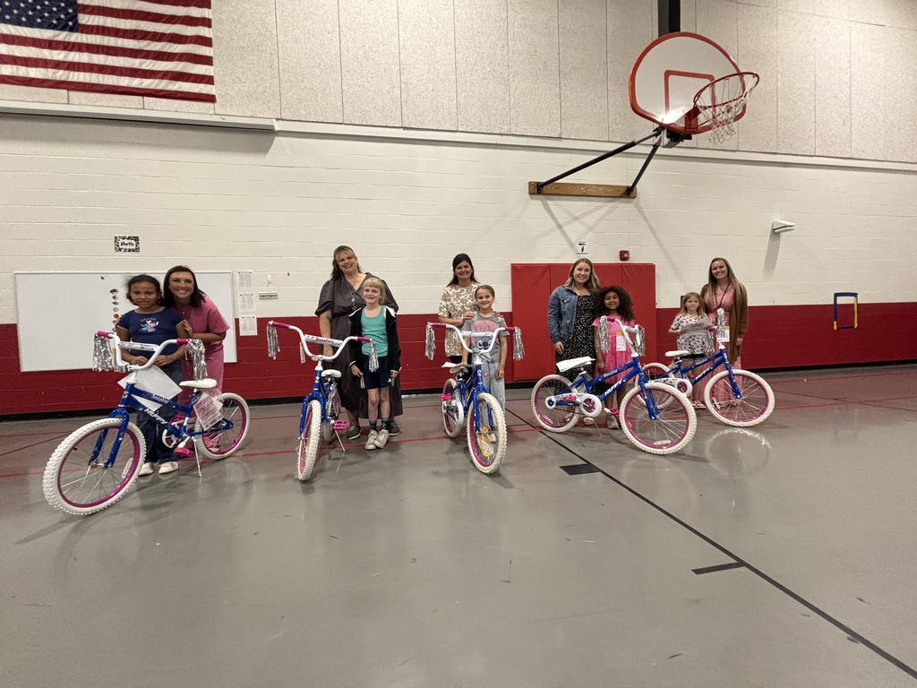 Group of children with bicycles in a gym. A basketball hoop and flag are in the background.