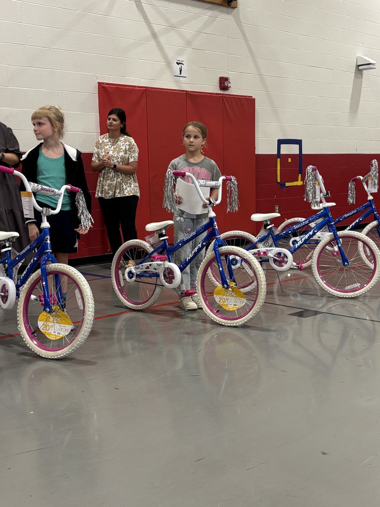 Girls stand beside bikes in a gym with a red wall and a white ceiling. Other adults stand behind them.