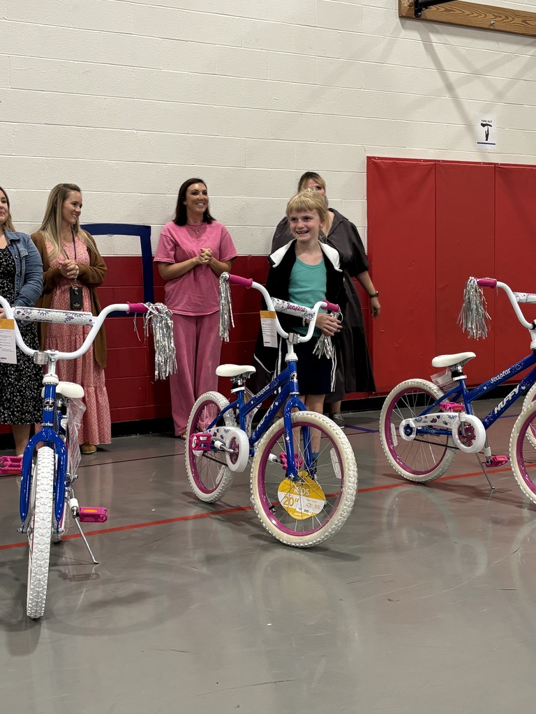 A young girl stands between two bicycles in a room with a red partition. Other adults stand behind the bicycles.
