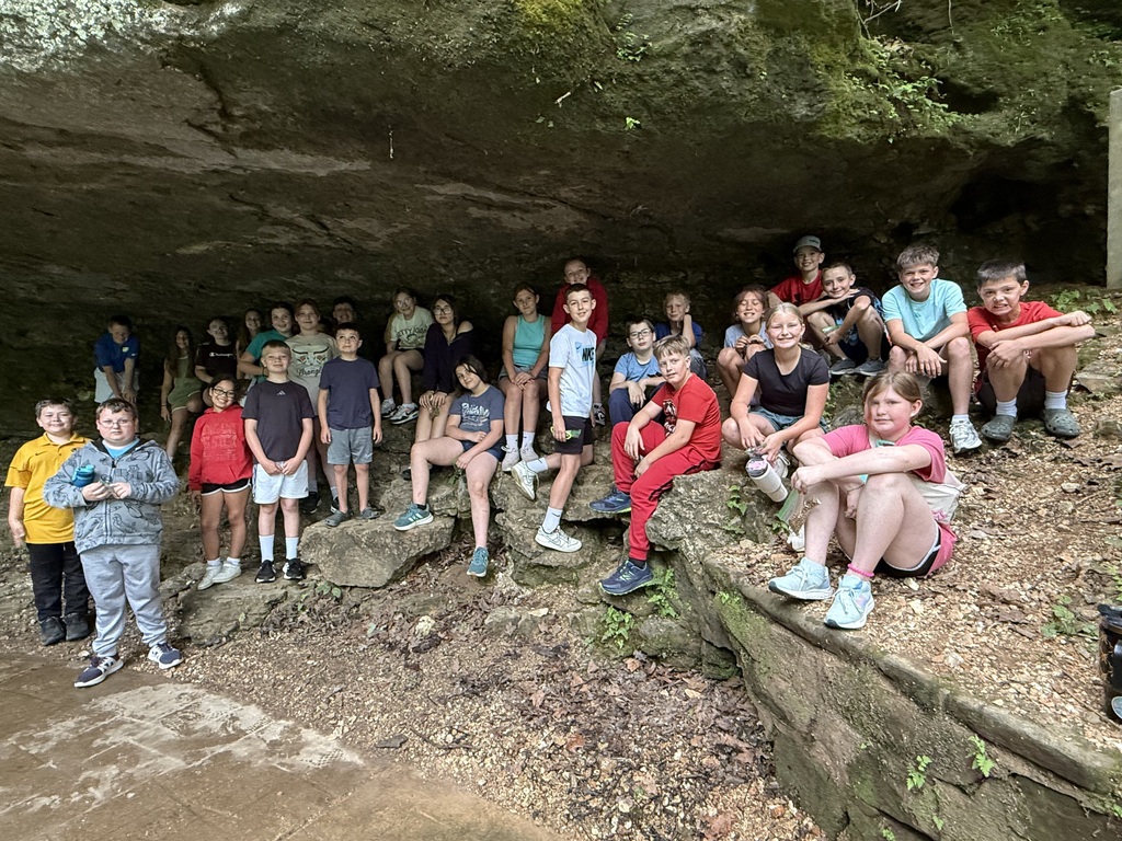 A group of children and adults are gathered around a cave entrance, sitting on rocks. Some wear hats.