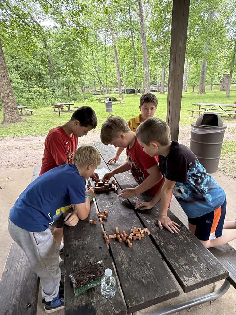 Five children work together at a picnic table. One is seated while the others lean over the table.