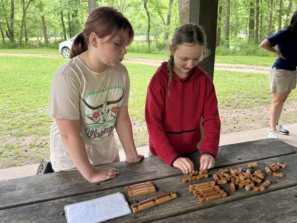 Two girls, one in white and one in red, work together at a table with wooden pieces. Behind them, another person stands near a car.