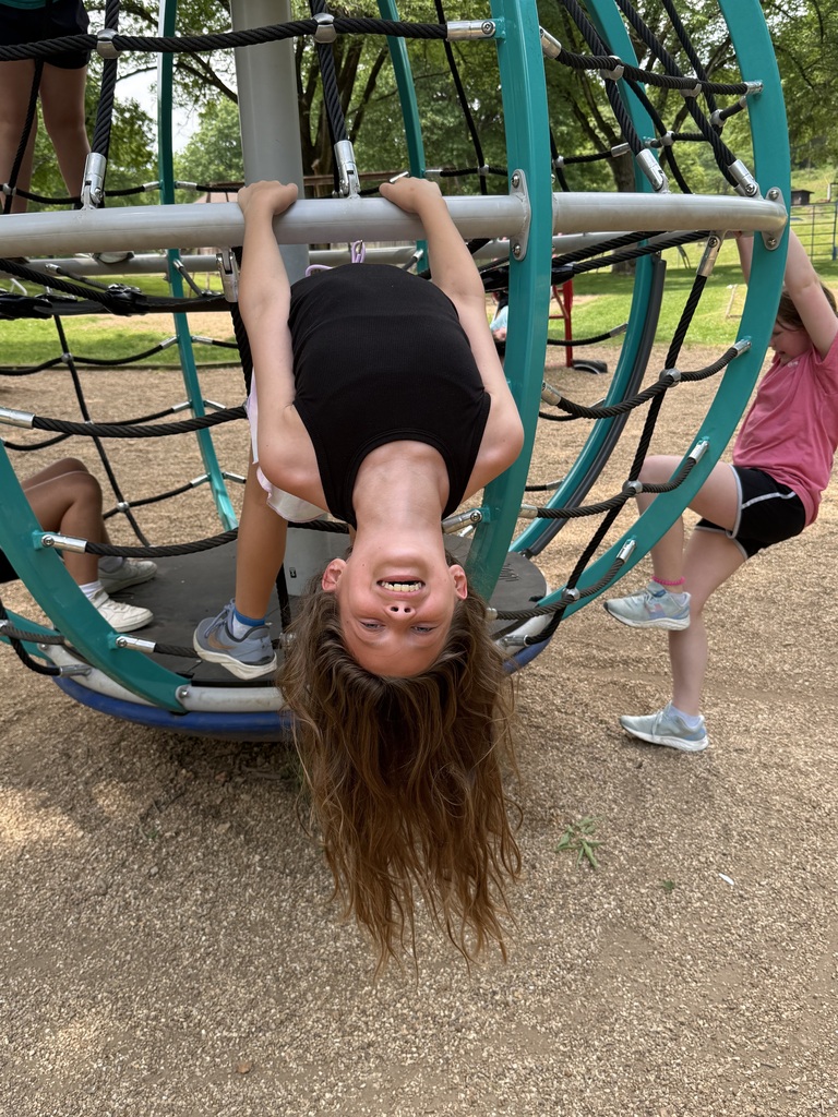 Children play on an outdoor climbing structure. One child hangs upside down, smiling. Another child is on the structure to the side.