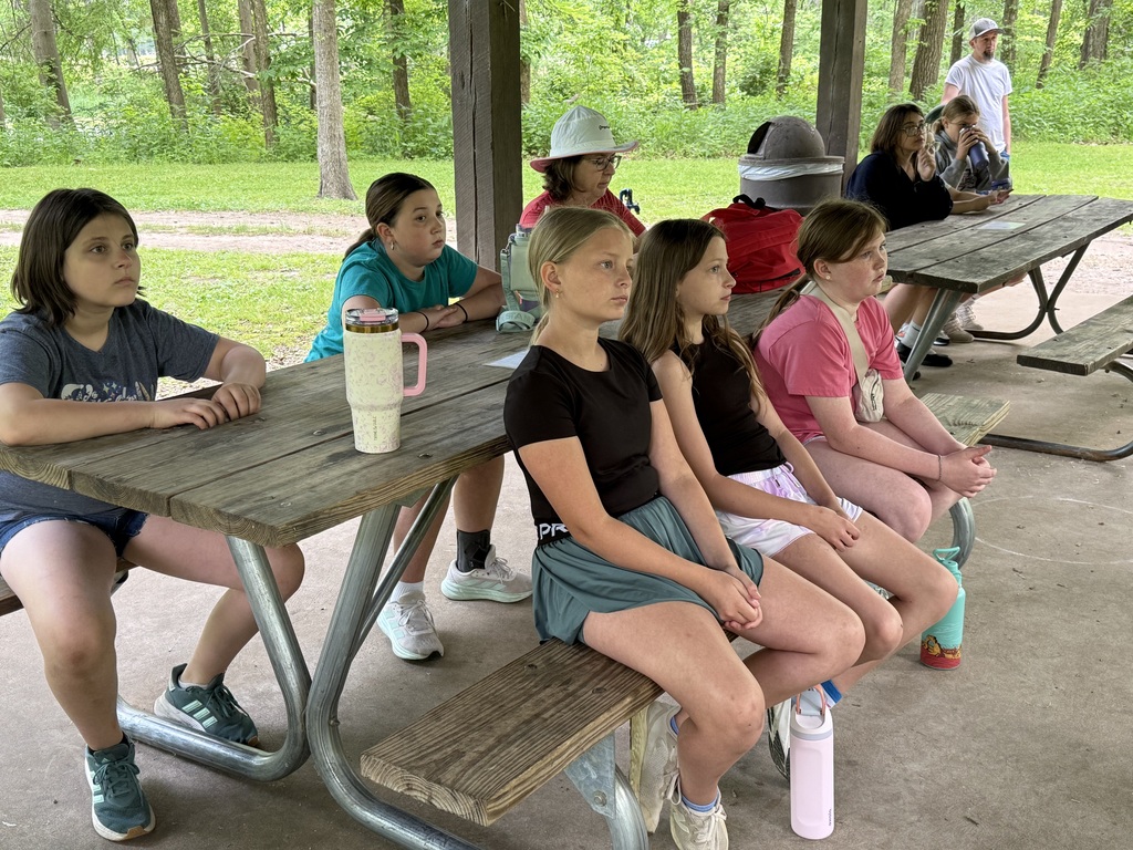 A group of girls sit at a picnic table, wearing casual clothing and sneakers. Behind them, people sit at another table.