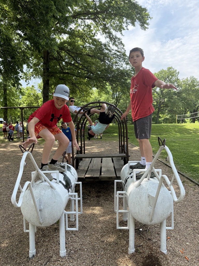 Two boys stand atop white and black figures at a playground, one holding a bar. Trees and a grassy area are behind them.