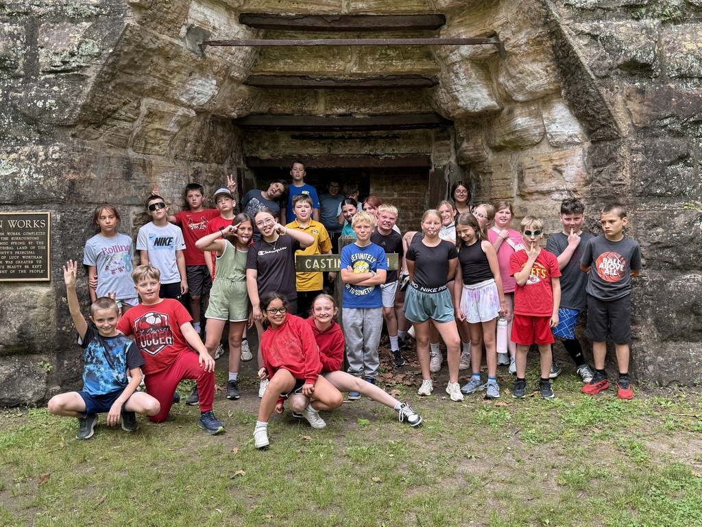 A group of individuals in casual clothing poses in front of an old stone archway. Some wear hats.
