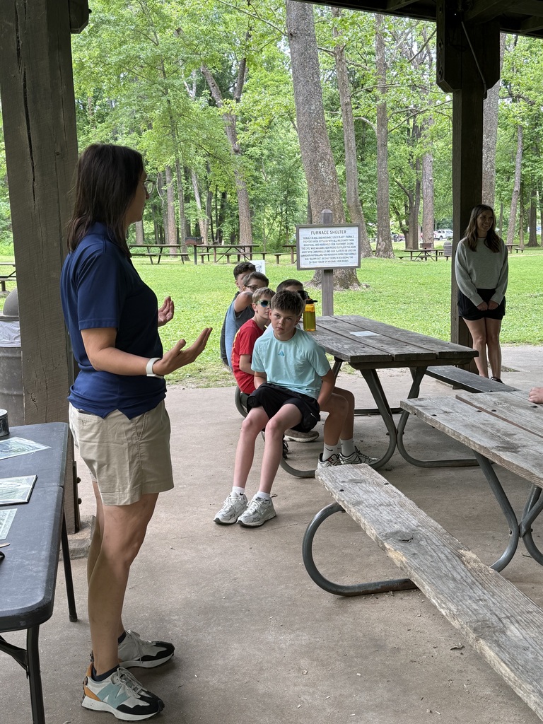 A woman addresses a group of children at a picnic table. The group sits on a bench. Trees and grass are in the background.