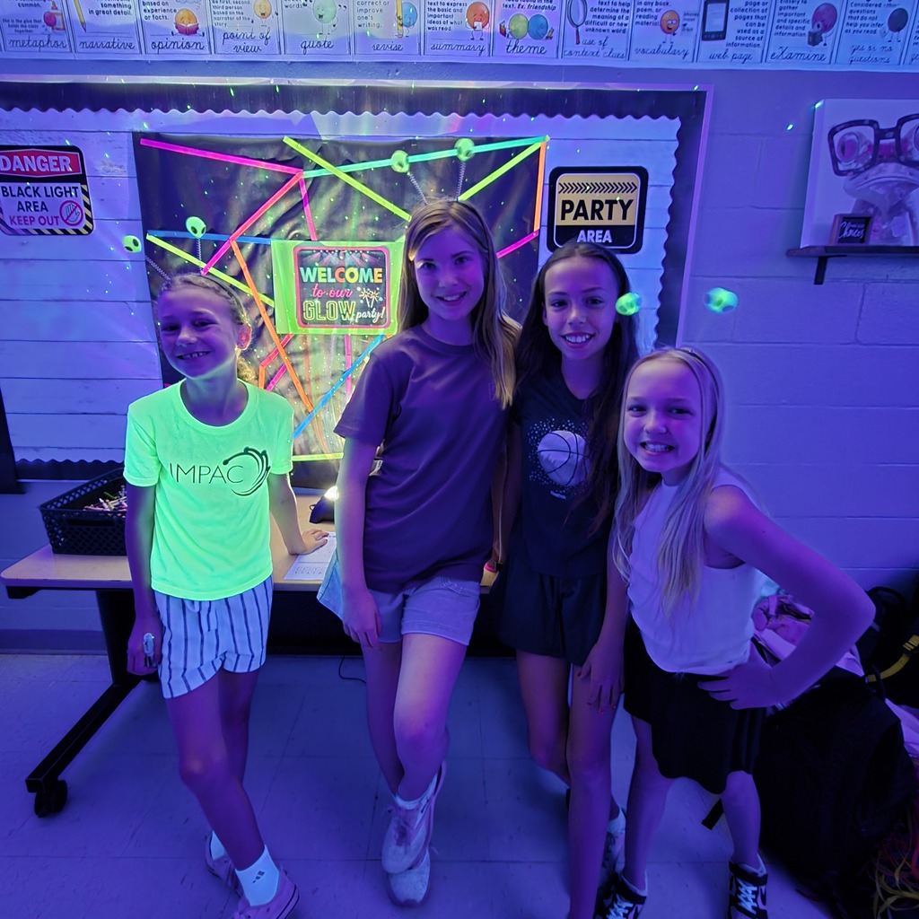 Four girls are standing in a room with a glowing purple background. One wears a green shirt and white shorts.