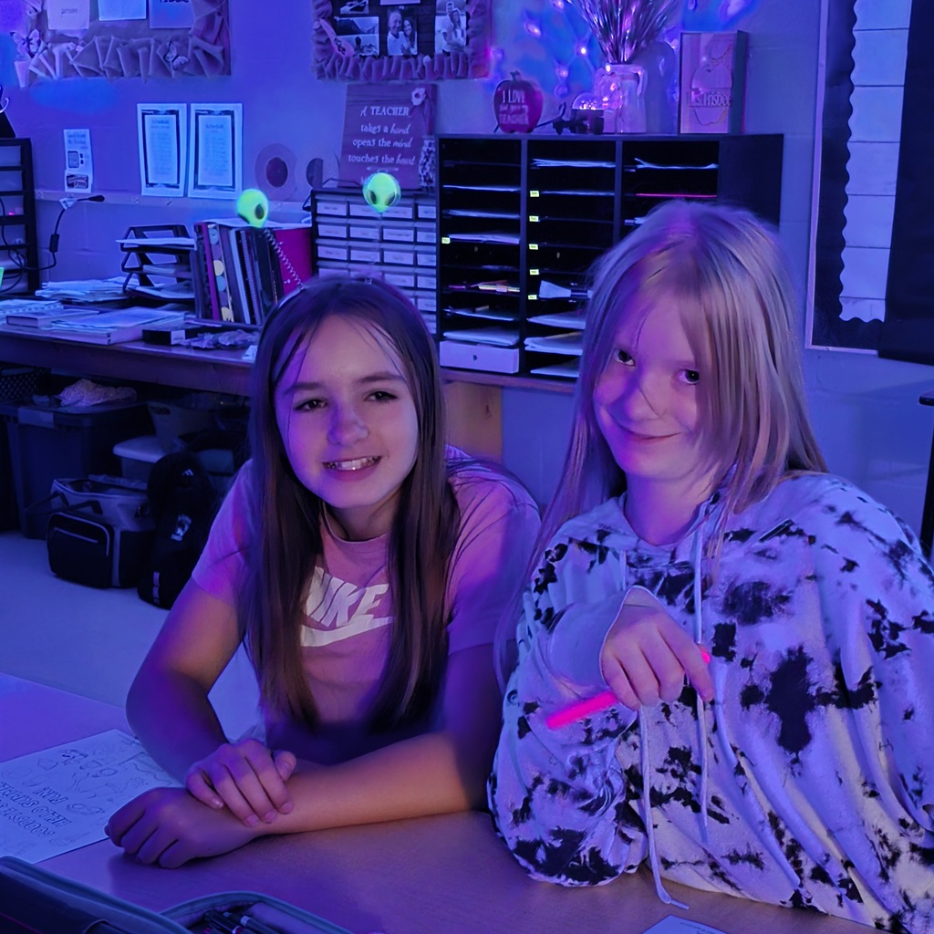 Two girls in a classroom with purple lighting, seated at a desk. One holds a pink object.