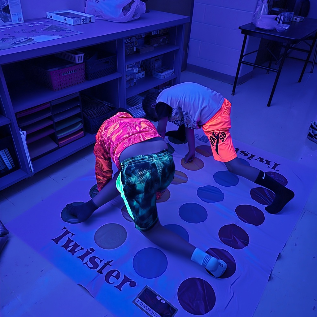 Two people play a game of Twister on a floor, wearing bright neon clothing. Shelves and tables in background.