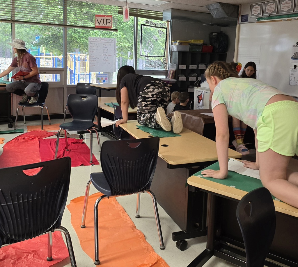 A classroom with desks and chairs, students doing exercises, colorful mats, a whiteboard, and a window.